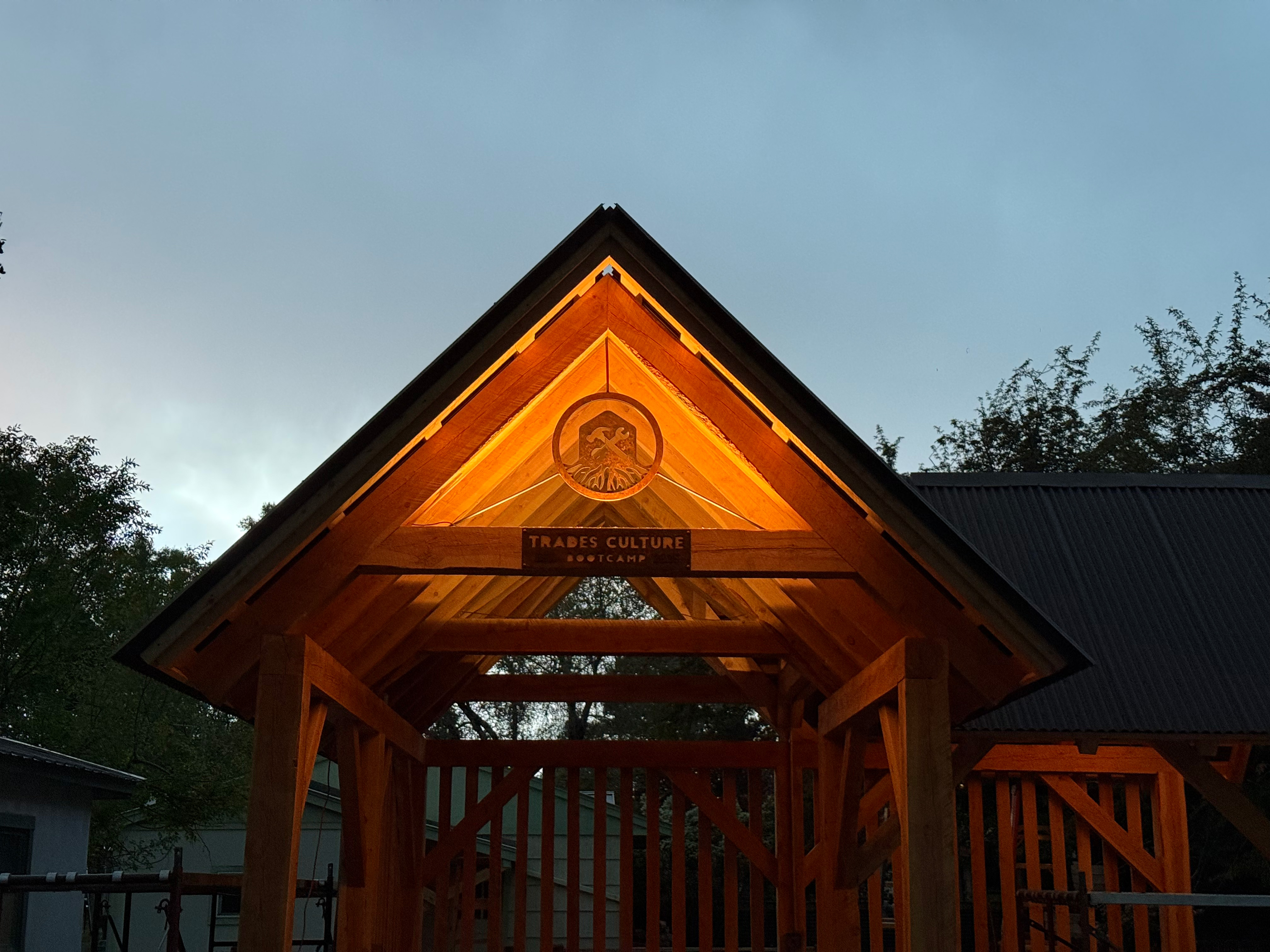 A photo of the solar powered gazebo lit up at night. Plaques created by the students, one reading “Trades Culture Bootcamp” and the other depicting a crossed hammer and wrench with roots radiating out below them, are visible in the peak of the structure. 